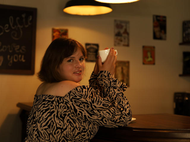 StaceyLex, a young woman with short brown hair, sits at a wooden table holding a white cup with both hands. She's wearing an off-shoulder, black and white zebra-patterned shirt. The warm, yellow overhead light casts a soft glow on her face. Her expression is slightly smiling. The background has several small, colorful framed pictures on a white wall. The image has a casual, cozy atmosphere with a focus on StaceyLex's relaxed, intimate moment. The lighting and composition emphasize her presence and the texture of her shirt.