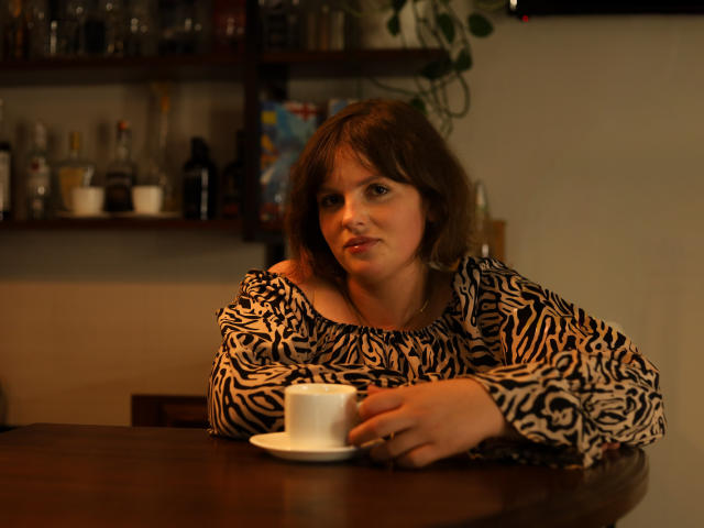 StaceyLex, a woman with medium-length brown hair, sits at a wooden table holding a white teacup on a saucer. She wears a black and white zebra-patterned blouse. Her expression is neutral, looking slightly to the left. The warm lighting casts soft shadows. The background includes a shelf with various glass bottles and plants. The image has a slightly blurred, realistic style. StaceyLex's right hand rests on the table while her left arm supports her head.