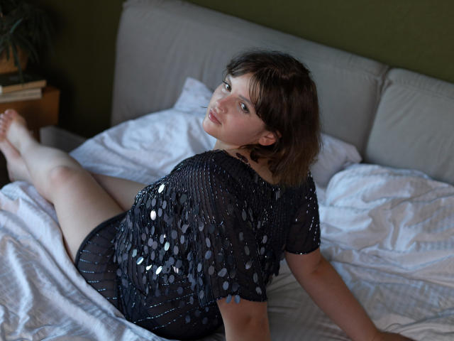 StaceyLex lies on a bed, her head turned to the camera with a slight smile. She has short brown hair and fair skin. She wears a black, sheer, sequined top with circular patterns, and black shorts. Her right arm is extended behind her, and her legs are stretched out. The white bed linens are slightly wrinkled. The green wall and wooden nightstand in the background are blurred. Natural light from the left. 