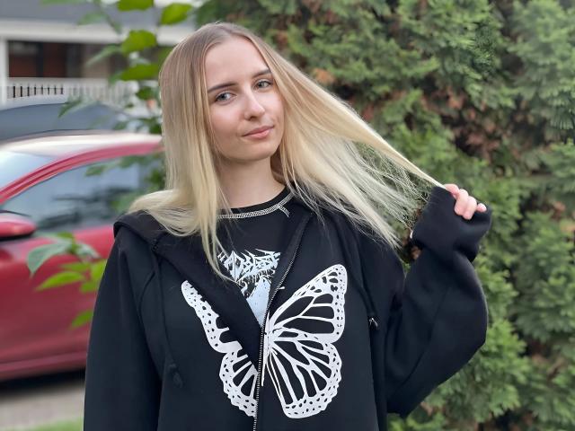 CutiCrystall-hot with long blonde hair, wearing a black sweatshirt with a white butterfly, holding her hair, standing in front of a red car and green bush.