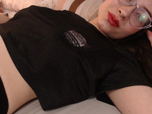 ElizabethMorrison lying on a bed, wearing a black t-shirt, glasses, and red lipstick, with her tits and black panties visible, her face showing a slightly bored expression.