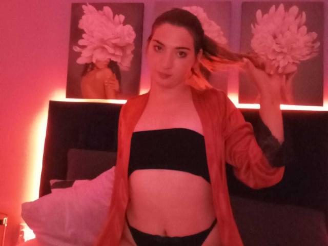 MedeyaStar with medium tits, black strapless top, red open shirt, black panties, holding her ponytail, red lighting, three pink flower paintings in background.
