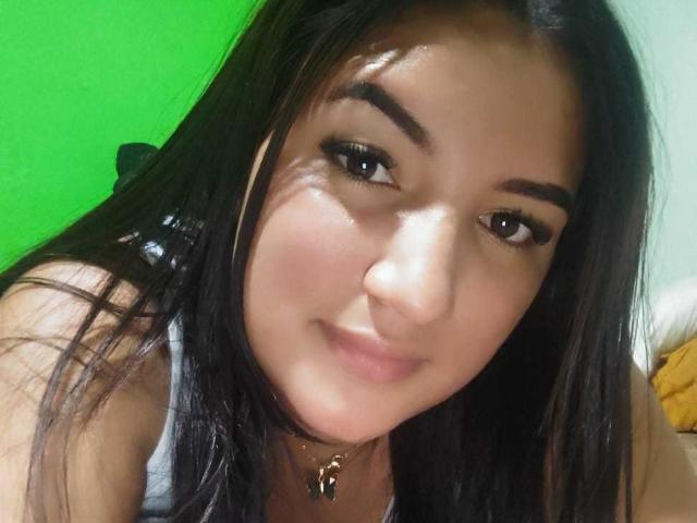 CharlotteVallie with long black hair, brown eyes, and a slight smile, wearing a gray tank top and a gold necklace, looking at the camera with a soft expression.
