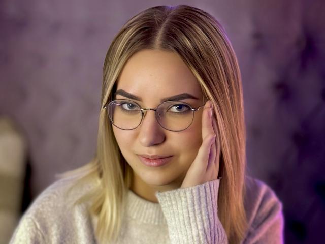 CarollFlex met blond haar, een bril en een witte trui, die haar gezicht aanraakt met een lichte glimlach. Haar borsten zijn niet zichtbaar, alleen haar gezicht is in beeld..
