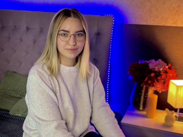 CarollFlex a les cheveux blonds, porte des lunettes et un pull rose clair, et est assise sur un lit éclairé par une lumière LED bleue. Ses fesses sont partiellement visibles, avec un oreiller derrière..