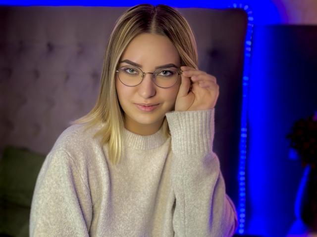 CarollFlex, con cabello rubio, gafas y un suéter blanco, tocándose la cara con una sonrisa sutil, con una luz LED azul en el fondo..
