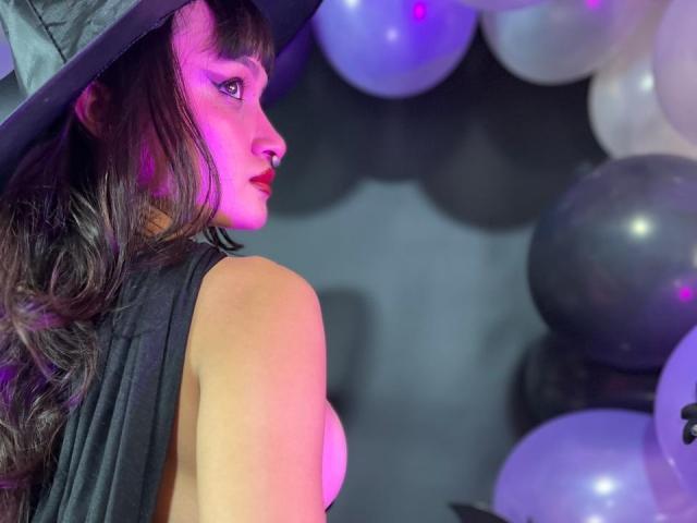 CandyyClair with wavy brown hair, wearing a witch hat, black dress, and purple lighting, showing her bare tits, surrounded by white and gray balloons.