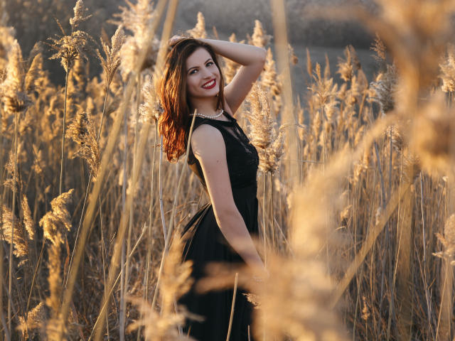AmyPure stands in a golden field, wearing a black dress, smiling with red lipstick, hand in wavy brown hair, tall grasses in focus, soft sunlight, sexy, confident.