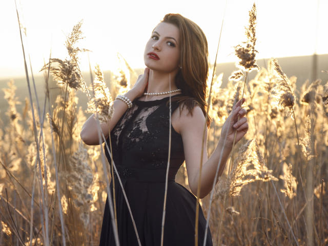 AmyPure, com cabelo castanho, vestindo um vestido preto de renda, segurando grama alta, usando uma pulseira de pérolas e um colar, em pé em um campo dourado, olhando para cima com batom vermelho..