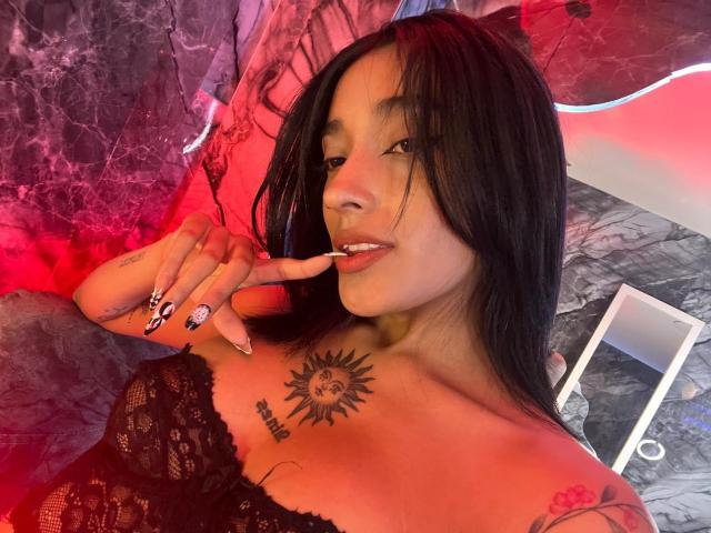 SalomeMille avec un haut en dentelle noire, des tatouages sur la poitrine et le bras, un doigt dans la bouche, de longs cheveux noirs, souriante, éclairage rouge et violet, montrant ses ongles peints, seins visibles..