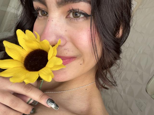 AitanaJohns with dark hair, green eyes, and a nose piercing, holding a yellow sunflower to her nose, showing off her painted nails, and a silver necklace.