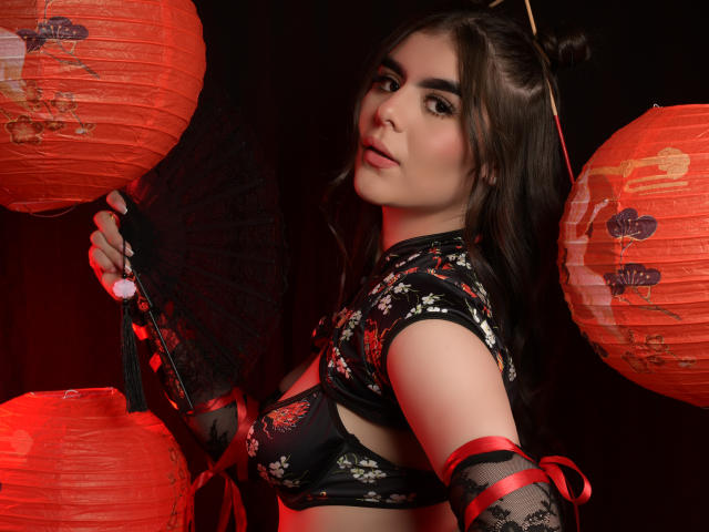 SofiaaFontaine with dark hair in a bun, wearing black floral top and red ribbons, holding black fan, surrounded by red lanterns, looking over shoulder with subtle smile.