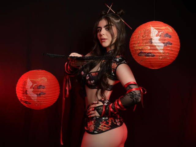 SofiaaFontaine with long brown hair in black floral lingerie, holding a black fan, red paper lanterns, and red ribbons, showing her ass and tits, in a dark room.