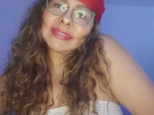 MiaAmoretti with curly brown hair, red beanie, glasses, and pink lipstick, showing off her tits in a white strapless top, smiling with glowing eyes.