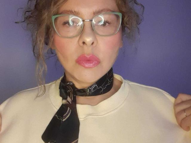 MiaAmoretti with curly brown hair, green glasses, pink lips, black choker, and white sweater, making a pouty face, holding sweater edges. Her tits are small and she's cute.