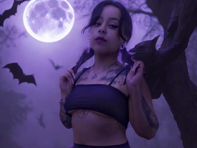 DaisyFx con trenzas, tatuajes y una blusa negra transparente, sosteniendo un bate, iluminada por la luz de la luna, con un brillo púrpura, murciélagos en el fondo, mirando hacia abajo con un piercing en la nariz y labios rosados..