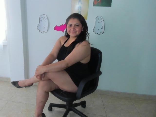 TiffanyAmazing sits in a black chair, wearing a black dress, with her legs crossed, showing her ass, and smiling. She has black hair and is in a light room.