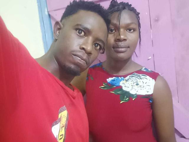 Photograph of LykyBlacks. A black man with short, curly hair and a thin beard stands next to a black woman with braided hair. The man wears a red shirt with a yellow graphic, while the woman wears a red floral dress. Both have neutral expressions. The woman's dress features large white and blue flowers. The man's left arm is around the woman's back. The woman's right arm is around his shoulder. The image is a close-up.