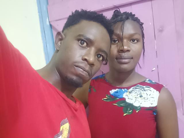 A photo of two LykyBlacks standing close together. The one on the left has short, curly black hair and a red shirt with a small logo. He looks directly at the camera with a neutral expression. The LykyBlack on the right has braided hair, wearing a red floral dress with a white rose pattern. Both have dark brown skin and are standing against a pink door. The image is a selfie with a slightly tilted perspective.