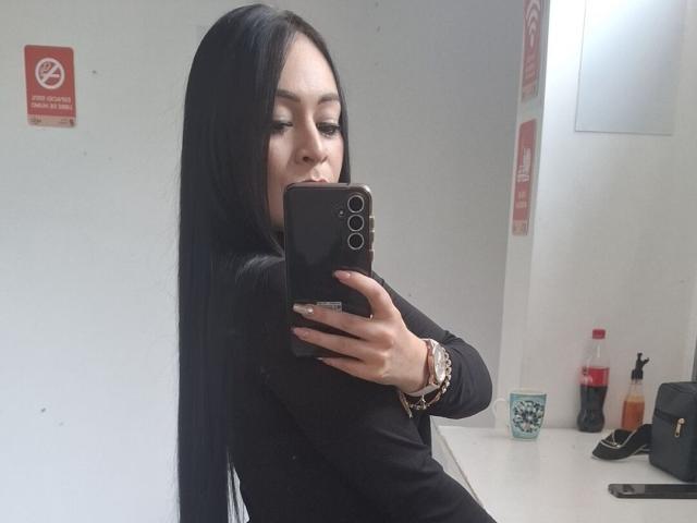 KristelRouse with long black hair, black long-sleeve shirt, holding phone, taking mirror selfie, pouty lips, bracelet, white wall, black bag, bottle, cup on white countertop.