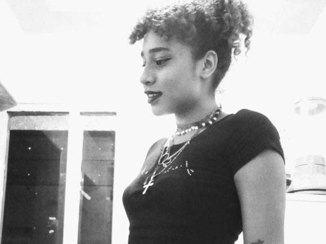 Black-and-white photo of NaomiMoss with curly hair, wearing a tight shirt, necklace with cross, and beaded choker. She's looking to the side with slightly parted lips.