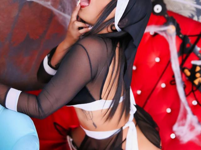 NenaBlack in a black sheer top and white lingerie, tongue out, hand near mouth, black hair, tattoo on side, red and blue background, black and white headwear.