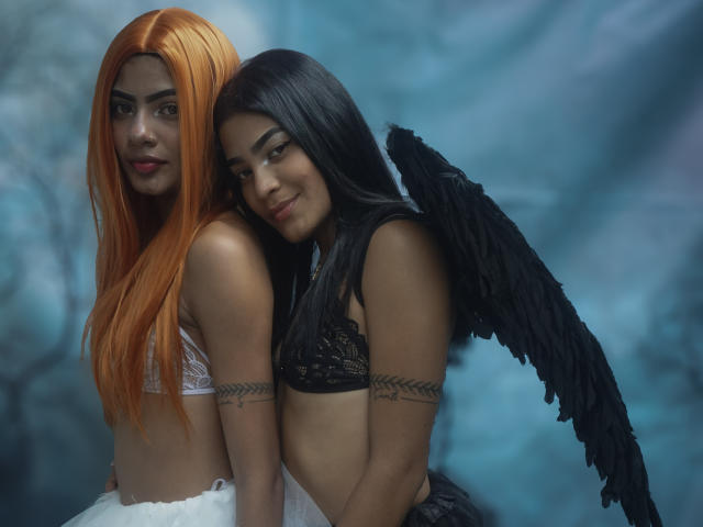 KarenAndLaura with orange hair and white lace bra, standing next to black-haired KarenAndLaura with black wings, lace bra, and arm tattoos, both showing tits and cute smiles.