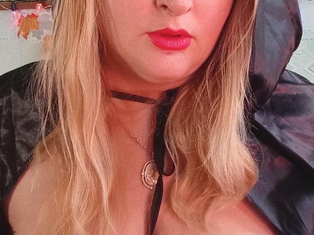 KelliRedhaer has blonde hair, red lipstick, a black choker with a pendant, and a black hooded garment revealing her cleavage and a black strap.