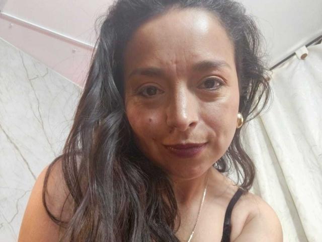 StangelarRivera with dark, wavy hair, brown eyes, and a small mole on her left cheek, wearing a black strap top, and a subtle smile, with purple lipstick.