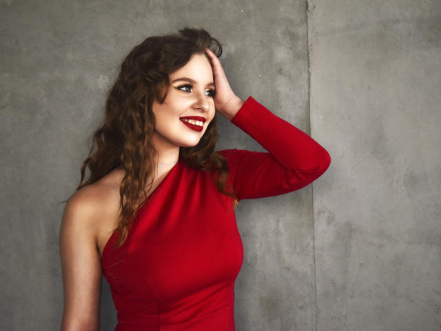 KyleJackson with wavy brown hair, red long-sleeve dress, hand on head, smiling with red lipstick, showing off her tits against a grey concrete wall.