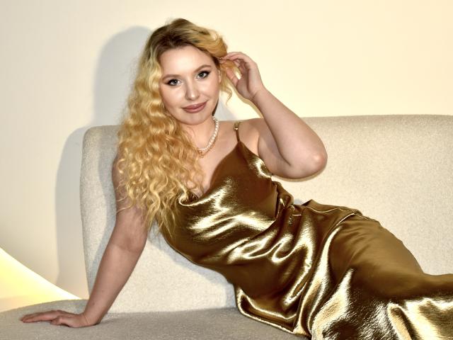 KyleJackson with long blonde curly hair, wearing a shiny gold dress, sitting on a beige couch, hand in hair, smiling, pearl necklace, pale skin, cute, sexy.