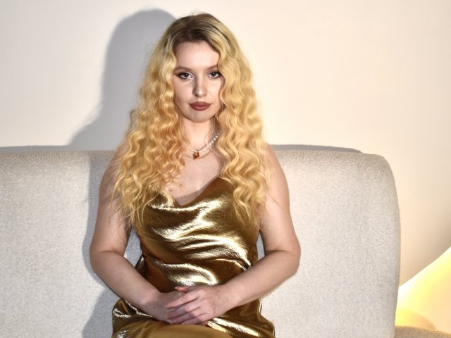 KyleJackson with long, wavy blonde hair wearing a shiny gold dress, sitting on a beige couch, hands in lap, looking at the camera with a serious expression.