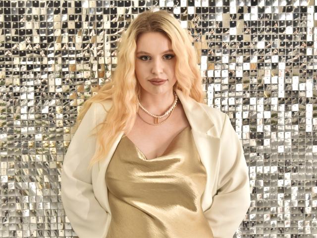 KyleJackson with blonde hair, wearing a shiny gold top, white jacket, and pearl necklace, has her hands in her pockets, standing in front of a metallic tile backdrop.