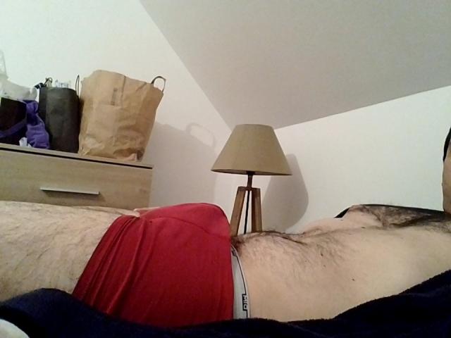 Paul69 liegt auf einem Bett, trägt rote Boxershorts, wobei sein behaarter Schambereich und ein weißer Bund sichtbar sind. Eine beigefarbene Lampe steht auf einer hölzernen Kommode..