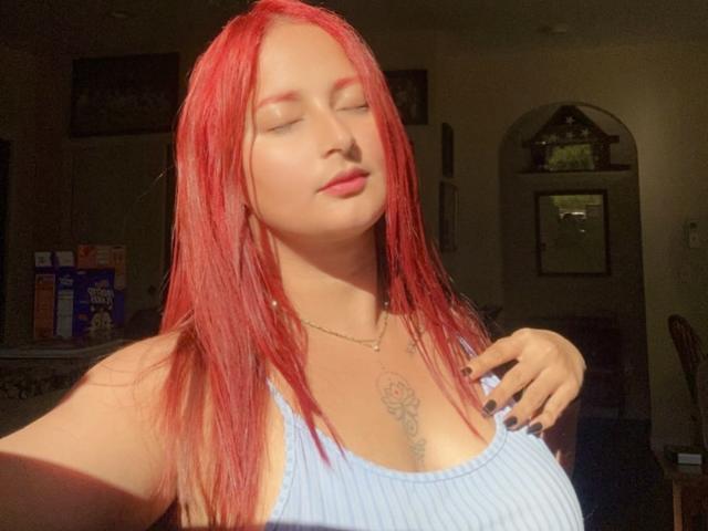 LatineMuse with red hair, eyes closed, wearing a blue ribbed tank top, hand on chest, showing her small tattoo, sunlight on her face, looking relaxed.