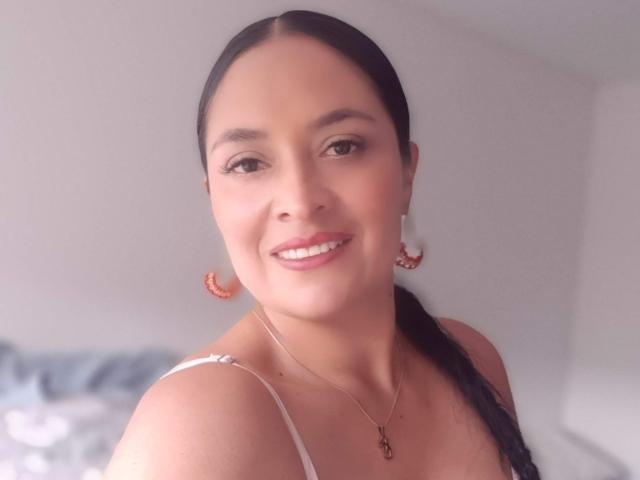 CatleyaSmith with black hair in a braid, smiling, wearing white straps, orange earrings, and a necklace, has a cute face, and is showing off her tits.