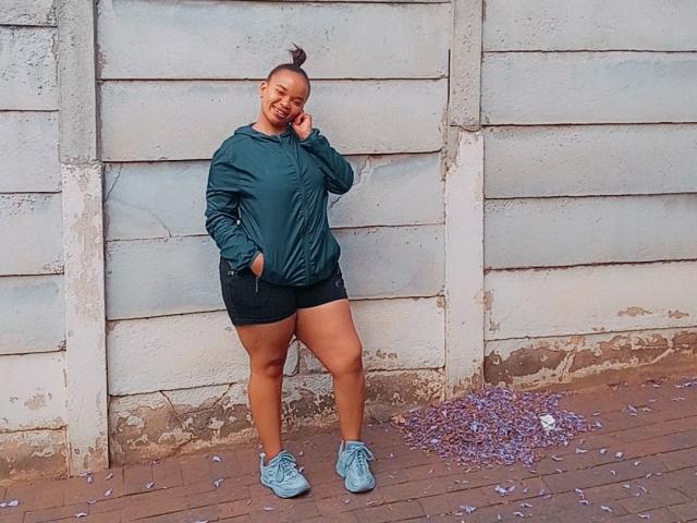 Mellorh6 stands against a worn, white, wooden wall. She wears a dark green hoodie, black shorts, and light blue sneakers. Her right hand is in her pocket, while her left hand touches her cheek. Her hair is in a high bun. The ground is brown with purple confetti. She has a relaxed, confident posture. The image is clear with natural light.