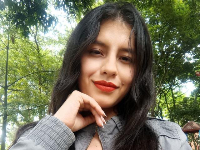MadissonFontaine with long black hair, orange lipstick, and gray jacket, smiling slightly, hand near face, green trees in background, sunlight filtering through leaves.