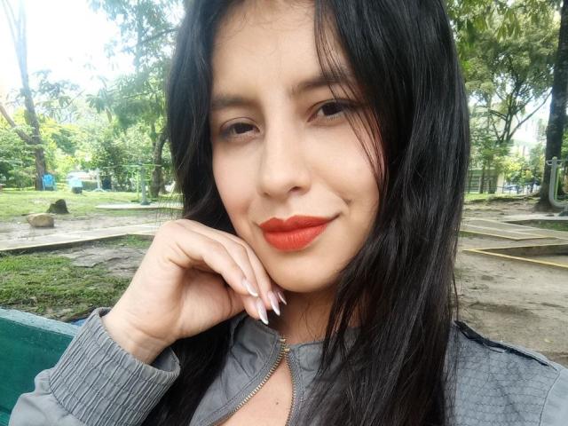 MadissonFontaine has black hair, red lipstick, and a grey zip-up jacket, with a hand resting on her cheek, smiling slightly in a park. Her ass is not visible.