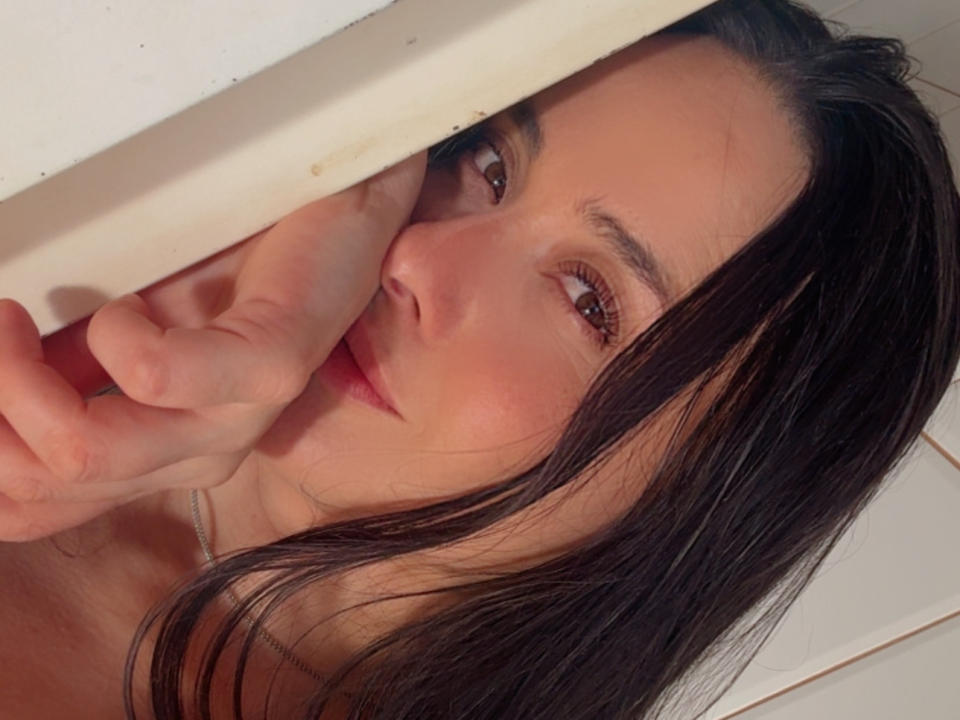 El rostro de Mia Alencar, con ojos marrones, cabello negro y una sonrisa sutil, sosteniendo su barbilla debajo de una viga de madera, mostrando sus labios rosados y un collar de plata..