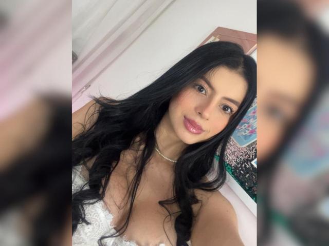 IsabellaJames with long black hair, shiny pink lips, white lace top, and a pearl necklace, smiling at the camera, showing her tits, in a selfie.