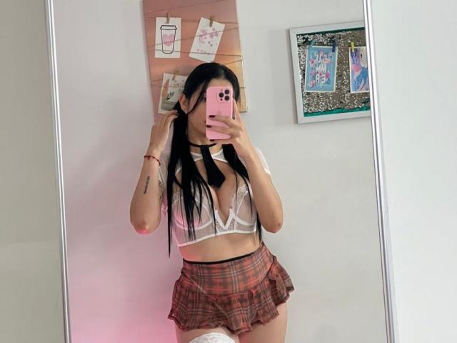 Isabella James macht ein Selfie vor einem Spiegel, auf dem sie ein durchsichtiges, weißes BH-Top mit sichtbaren Brustwarzen, einen roten karierten Minirock und weiße, hohe Socken trägt. Sie hält ein rosa Handy..