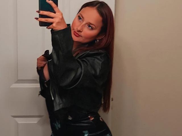 LatineMuse met lang rood haar, een zwarte leren jas en donkere broek, die een spiegelselfie maakt, een groene telefoon vasthoudt en met een lichte glimlach naar de camera kijkt..
