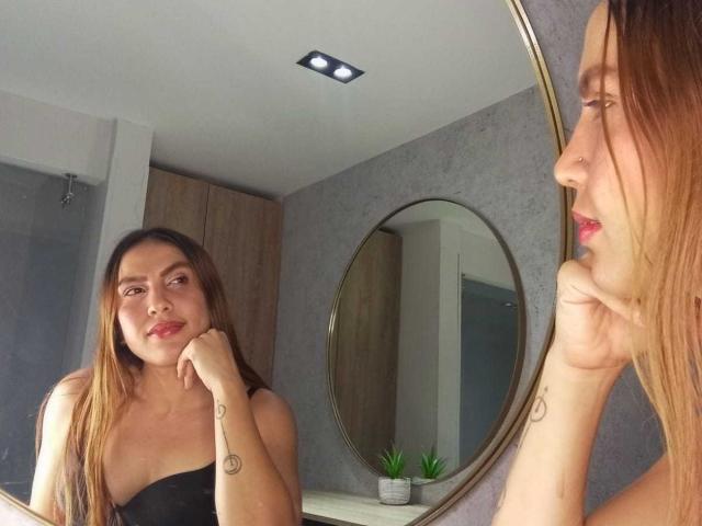 SusanThomass with long brown hair, nose ring, and small tattoo on wrist, stands in front of a mirror, looking at herself with a slight smile, wearing a black strapless top.