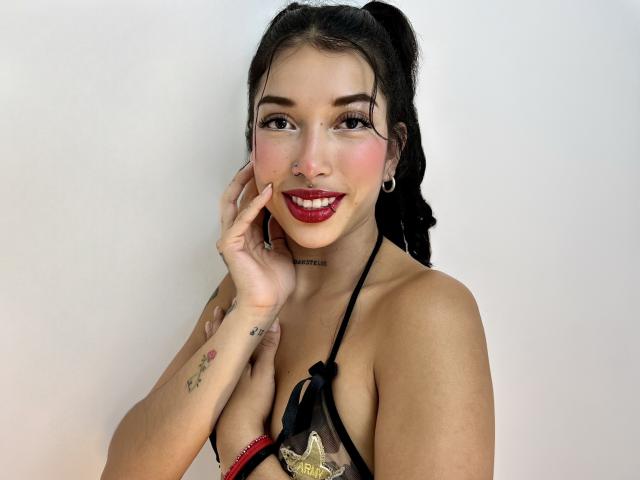KrystalEvans with dark hair in puffs, red lipstick, nose ring, black halter top, visible tits, tattoos, holding hand near face, smiling, white background.