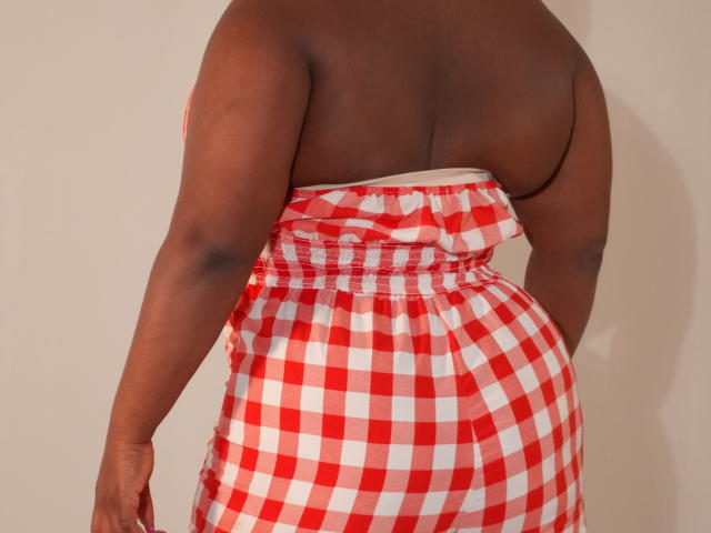Dilay's back shows dark brown skin and a red-white checkered dress with a low back, highlighting her ass and bare shoulders. Her arms hang by her sides.