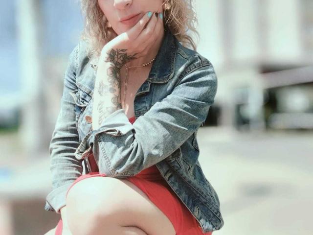 SammyDesire with wavy blonde hair, denim jacket, red dress, tattoo on right hand, hand on cheek, sitting, sunny outdoor background, slightly blurred. Her tits are subtle under the jacket.