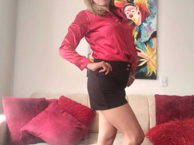 SammyDesire in a shiny pink blouse, black high-waisted skirt, showing her ass, standing with one hand on her hip, red pillows on a beige sofa.