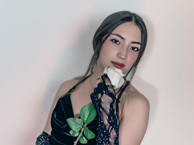 AnnFisher with long black hair, wearing a black lace glove, holding a white rose to her mouth, looking seductively at the camera, showing off her tits and a green leaf.