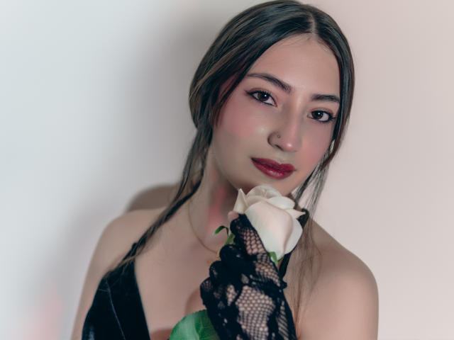Ann Fisher mit langem, braunen Haar, rotem Lippenstift, die eine weiße Rose hält und einen schwarzen Spitzenhandschuh trägt, blickt mit einem leichten Lächeln in die Kamera. Sie trägt ein schwarzes Top ohne Träger..
